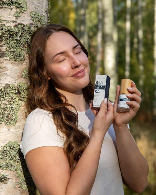 A woman standing by a birch tree in a sunny forest, smiling with her eyes closed while holding Saaren Taika Bladderwrack Super Serum. She appears relaxed and content, enjoying the warmth and calm of Nordic nature.